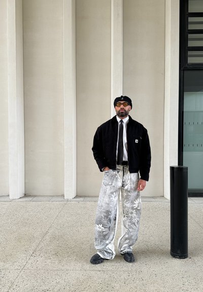 Chaqueta negra, camisa blanca y corbata, pantalones oversized grises con patrón floral, boina negra, gafas de sol amarillas, zapatos negros. Fondo de concreto.