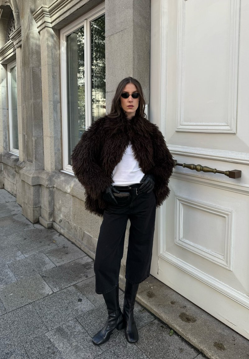 Femme portant une veste noire en fourrure, une chemise blanche, un pantalon noir, des bottes, des gants en cuir et des lunettes de soleil, se tenant contre un bâtiment en pierre et une porte blanche.