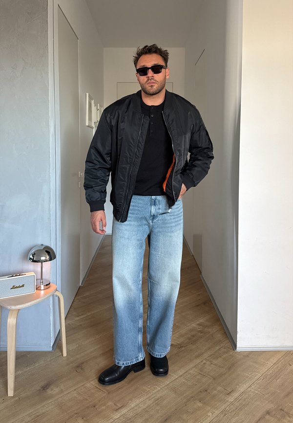 Un hombre posa en un pasillo con una cazadora bomber negra, una camisa negra, unos vaqueros azul claro, gafas de sol y botas negras.
