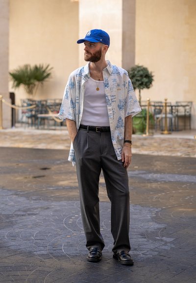 Camisa blanca de manga corta con un patrón azul, combinada con pantalones grises, una camiseta sin mangas blanca y una gorra de béisbol azul. Zapatos negros completan el conjunto.
