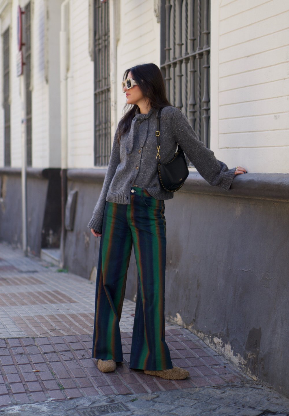 Cárdigan de punto gris con botones, pantalones de pierna ancha a rayas verdes y azules, y zapatillas de casa de pelo color beige; complementado con un bolso negro y gafas de sol.