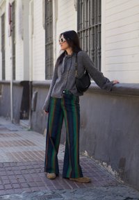Cárdigan de punto gris con botones, pantalones de pierna ancha a rayas verdes y azules, y zapatillas de casa de pelo color beige; complementado con un bolso negro y gafas de sol.