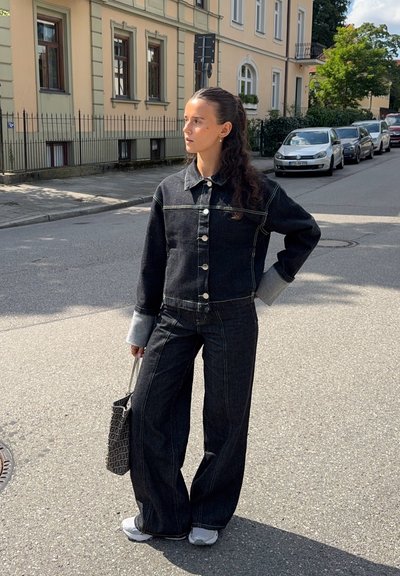 Chaqueta de mezclilla negra con puños en gris claro, cierre de botones; pantalones vaqueros de cintura alta y pierna ancha a juego. Lleva un bolso negro texturizado y calza zapatillas deportivas.