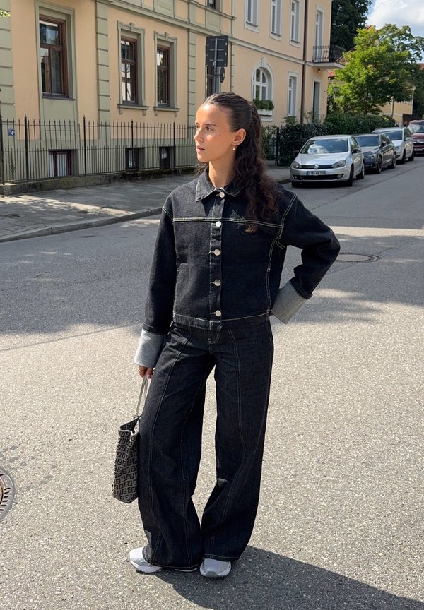 Black denim jacket with light grey cuffs and button front; matching high-waisted wide-leg jeans. Carrying a textured black bag; wearing sneakers.