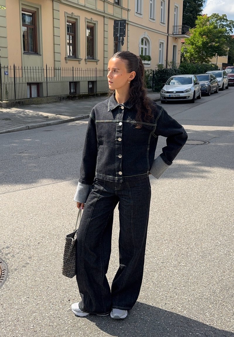 Veste en denim noire avec des poignets gris clair, fermeture à boutons ; jean large taille haute assorti. Porte un sac noir texturé ; chaussures de sport.