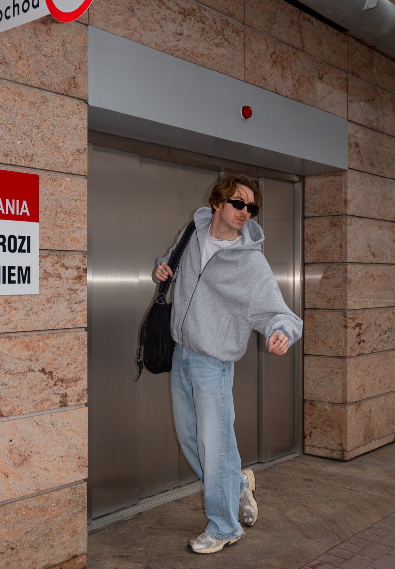 Junger Mann in grauer Kapuzenjacke, Sonnenbrille und Jeans steigt aus einem Edelstahlaufzug in einem Gang mit Steinfliesen eines Gebäudes aus und trägt eine schwarze Tasche.