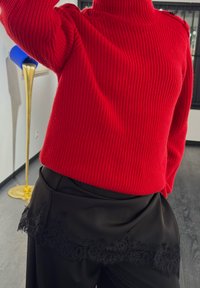 Gruby czerwony sweter z prążkowanym splotem i wysokim kołnierzem, zestawiony z czarnymi spodniami ozdobionymi koronkowym wykończeniem u dołu. Gładkie faktury i dopasowany krój.