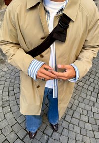 Osoba w beżowym płaszczu, pod którym ma niebiesko-białą koszulę w paski, białą koszulkę, niebieskie dżinsy i brązowe buty, stojąca na brukowanej nawierzchni.