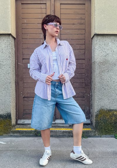 Camisa de rayas, de color púrpura claro y oversize con un bolsillo, camiseta blanca sin mangas, pantalones cortos de mezclilla azules, calcetines blancos y zapatillas blancas con detalles marrones.