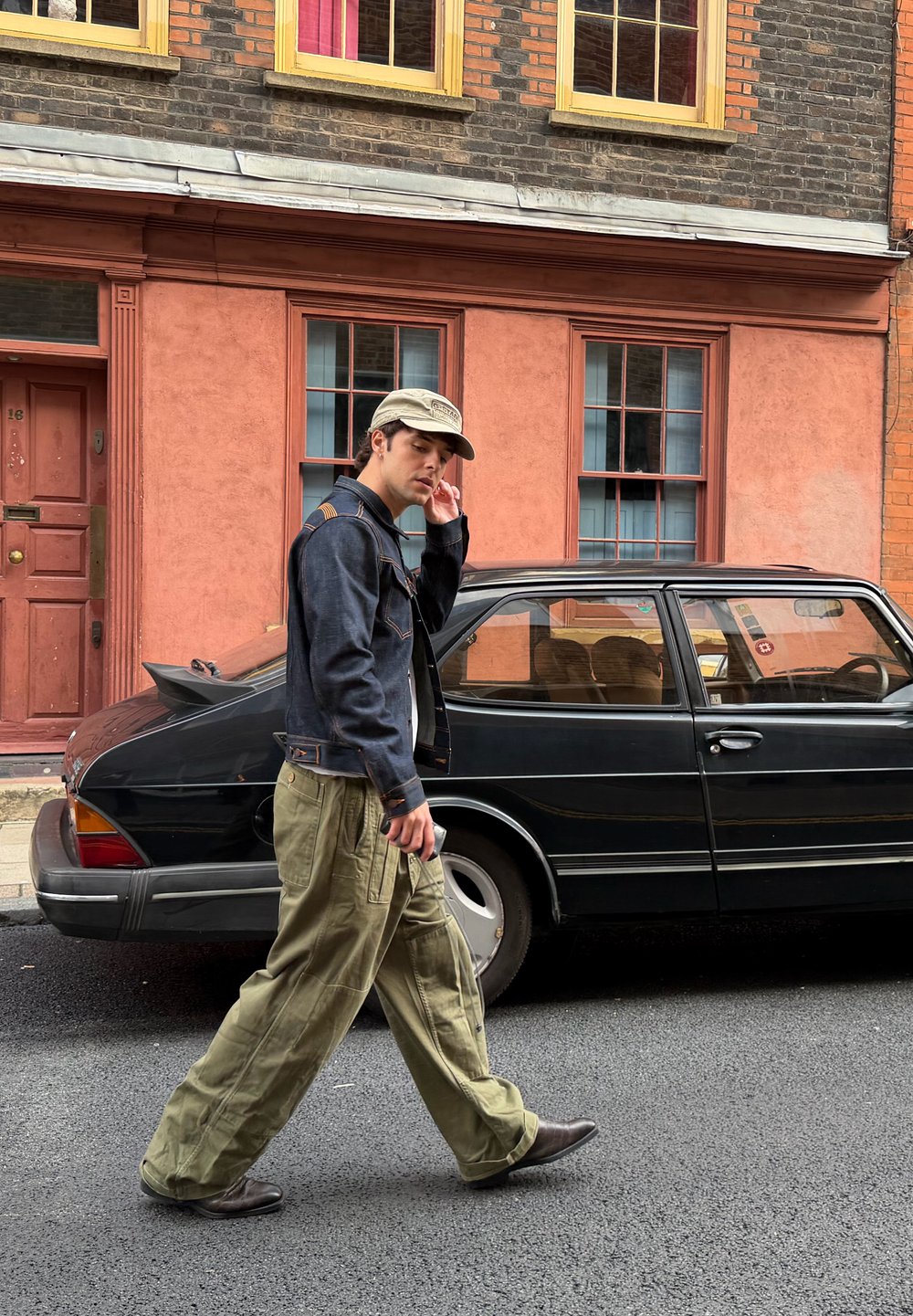 Spijkerjack met oranje stiksels, olijfgroene cargobroek en grijze pet gedragen door een persoon die langs een zwarte auto op een stedelijke straat loopt.