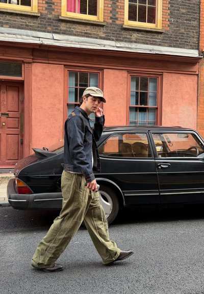 Chaqueta vaquera con costuras naranjas, pantalones cargo oliva y gorra gris llevados por una persona que camina junto a un coche negro en una calle de la ciudad.