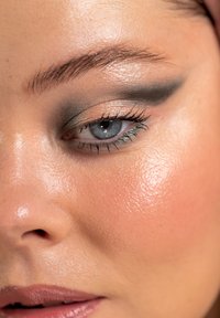 Close-up van een gezicht met blauwe ogen, verlichte huid, donkere oogschaduw en groene eyeliner. Gladde textuur en natuurlijke lipkleur zijn zichtbaar.