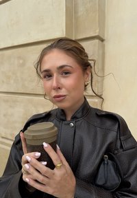 Giacca di pelle nera con texture e dettagli a bottoni a pressione, tiene in mano una tazza di caffè marrone. Orecchini a cerchio dorati e anelli. Unghie di un rosa chiaro.