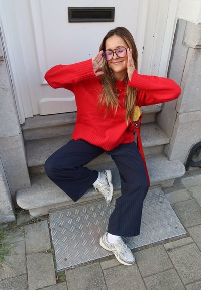 Jersey rojo de lana, pantalones anchos de rayas navy y zapatillas blancas. De pie en unos escalones con una placa de metal texturizada, con una puerta de fondo.