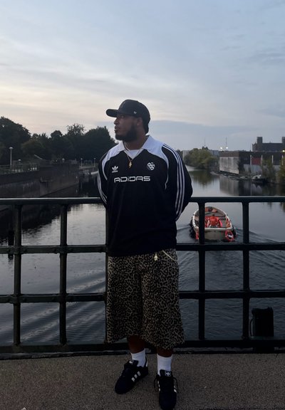 Jersey de Adidas en blanco y negro, pantalones cortos de leopardo, zapatillas negras, de pie junto a una vía fluvial, con un barco al fondo.