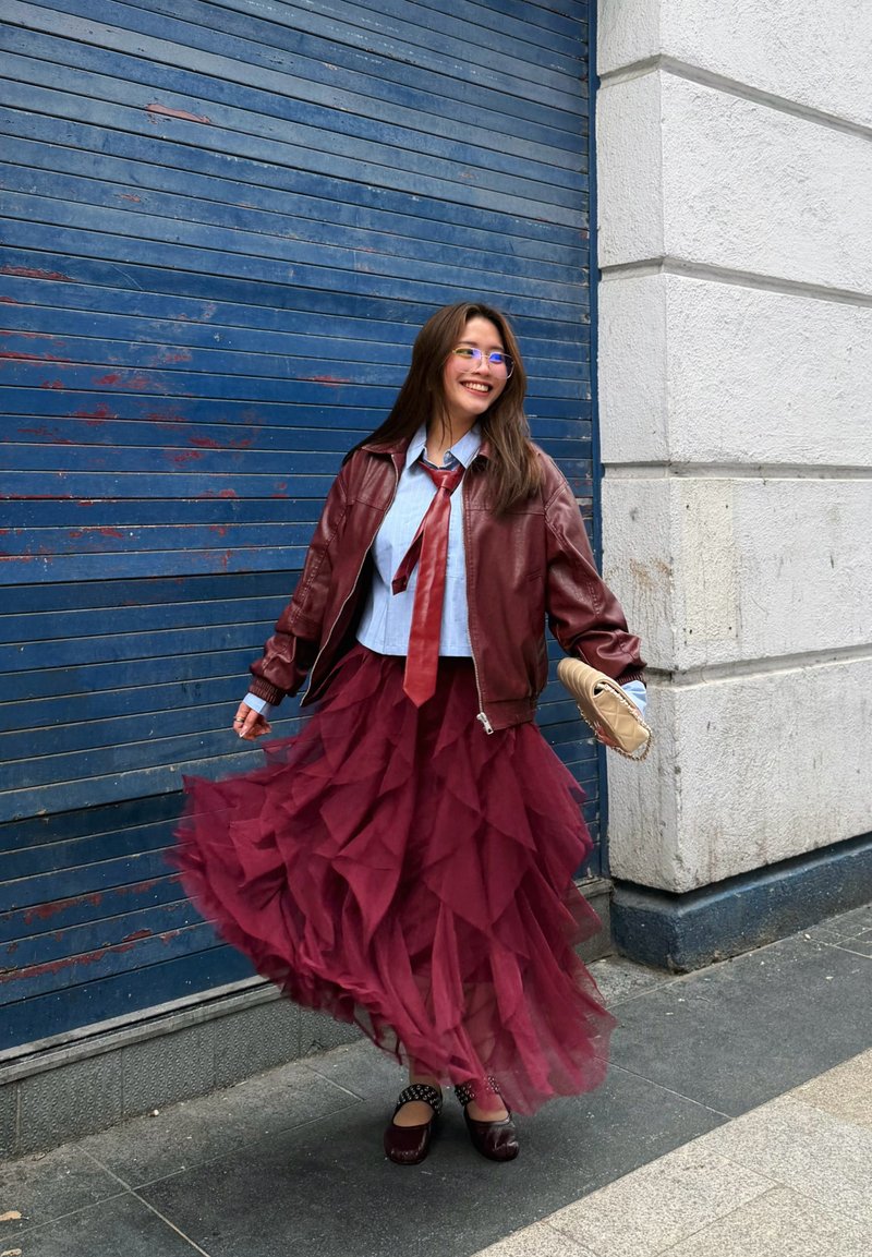 Frau lächelt und dreht sich in einem mehrlagigen bordeauxroten Rock, blauem Hemd, roter Krawatte, dunkelroter Lederjacke, hält eine beigefarbene Clutch vor einer blau geklappten Fensterlädenwand.