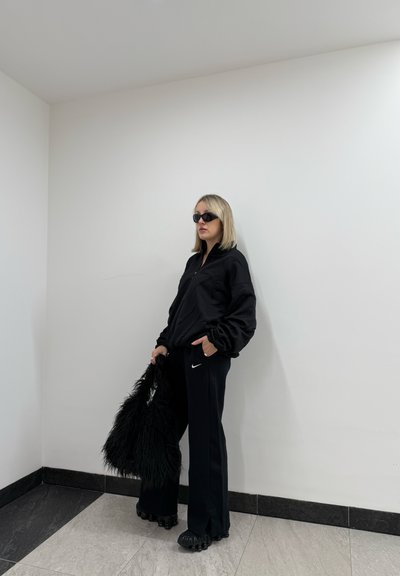 Chaqueta negra con cremallera y mangas abullonadas, pantalones deportivos negros con un logo blanco de Nike y un bolso negro esponjoso. Se llevan gafas de sol negras.