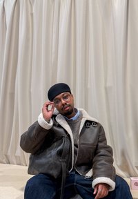 Chaqueta de cuero marrón con cuello de borrego, suéter gris debajo, camisa azul, vaqueros oscuros, sentado frente a un fondo claro con cortinas.