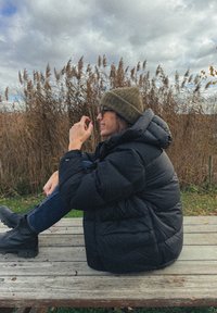 Γυναίκα με μαύρο μπουφάν τύπου puffer, πράσινο σκουφάκι και μπότες, καθισμένη σε ξύλινο παγκάκι σε εξωτερικό χώρο, με ψηλά χόρτα και συννεφιασμένο ουρανό στο φόντο.