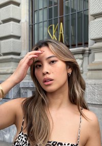 Jonge vrouw met lang, golvend haar, gekleed in een jurk met luipaardprint. Ze heeft een gouden armband en grote oorbellen, met een gebouw op de achtergrond.