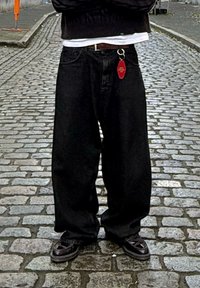 Jeans negros holgados con un llavero en el bolsillo delantero, llevados con una camiseta blanca y un suéter oscuro, complementados por unos zapatos oscuros con patrón.