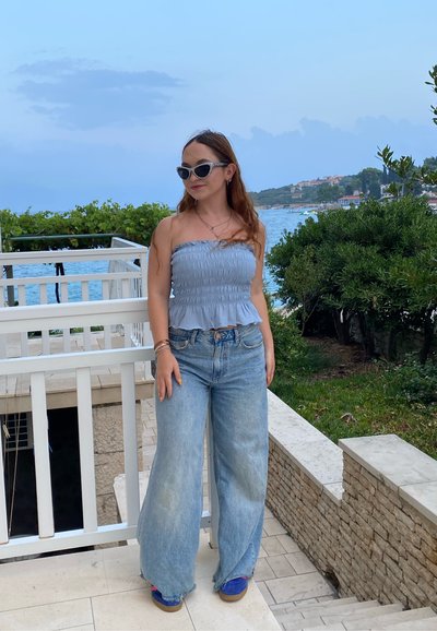Top sin mangas de color azul claro con volantes; jeans de mezclilla de tiro alto y pierna ancha de color claro; gafas de sol; calzado informal; entorno al aire libre con vegetación.