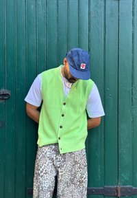 Πράσινο πλεκτό γιλέκο με κουμπιά πάνω από ένα άσπρο t-shirt, συνδυασμένο με ανοιχτόχρωμο παντελόνι με λεοπάρ σχέδιο και μπλε καπέλο σε πράσινο φόντο.