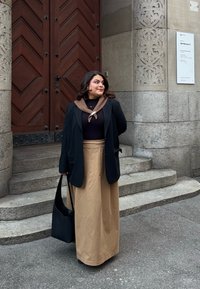 Sort blazer over en mørk bluse med en brun krave, parret med en beige maxinederdel. Holder en sort skuldertaske ved en stenstige.
