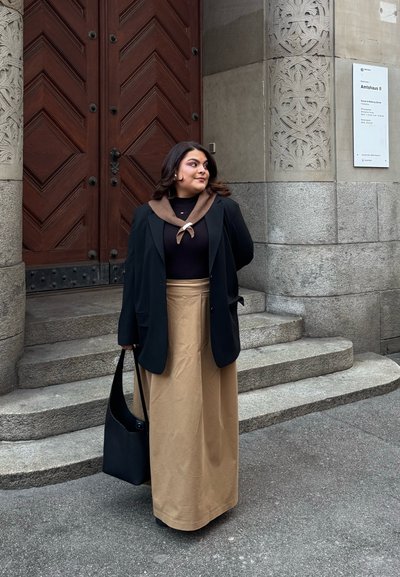 Blazer negro sobre una blusa oscura con cuello marrón, combinado con una falda maxi beige. Sostiene un bolso negro de hombro al lado de una escalera de piedra.