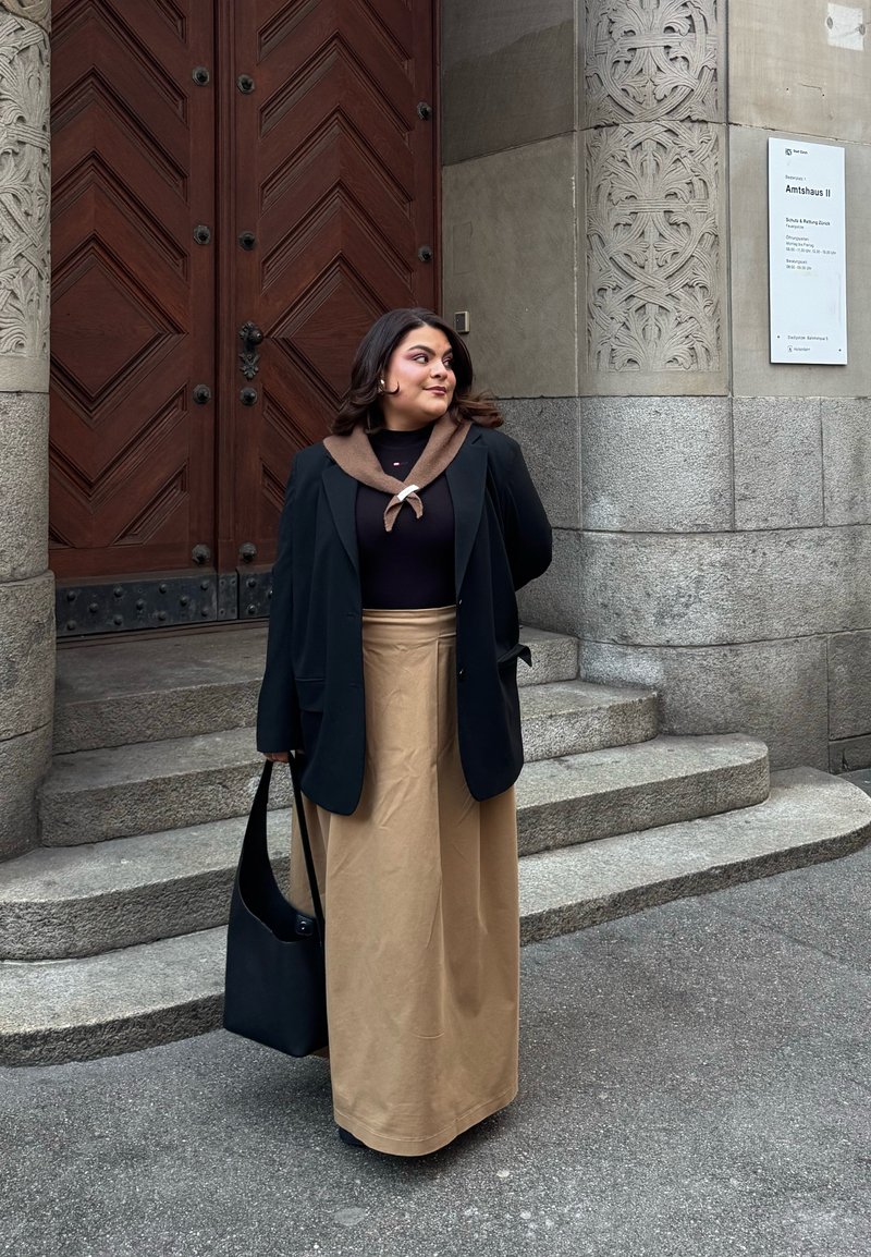 Zwarte blazer over een donker top met een bruine kraag, gecombineerd met een beige maxi rok. Houdt een zwarte schoudertas vast bij een stenen trap.