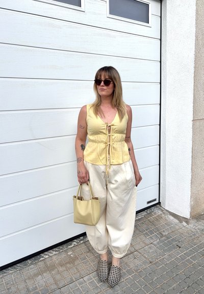 Mujer con top amarillo sin mangas, pantalones anchos color crema, zapatos sin cordones con estampado, gafas de sol y sosteniendo un bolso amarillo junto a una puerta de garaje blanca.