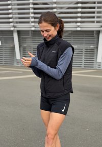 Czarna, ocieplana kamizelka z wysokim kołnierzem założona na niebieską koszulę z długim rękawem, czarne szorty oraz logo Nike. Sceneria na świeżym powietrzu z szarym chodnikiem.
