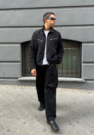 Chaqueta de tela negra con cremalleras plateadas, combinada con pantalones negros holgados y botas negras. Pared y pavimento grises de fondo.