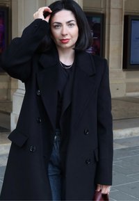 Zwarte dubbelgeknoopte jas met grote knopen, gedragen over een zwart topje en blauwe spijkerbroek. Houdt een kastanjebruine handtas vast. Steil zwart haar, lichte make-up.