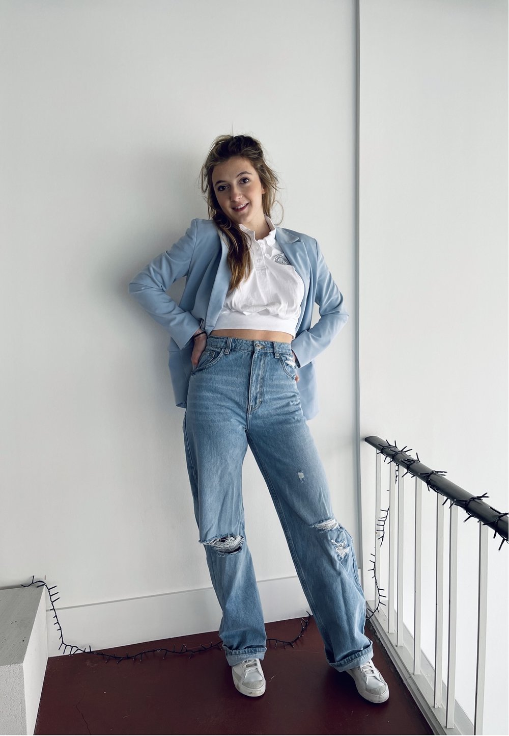 Lichtblauwe blazer over een wit cropped shirt, gecombineerd met distressed lichte jeans en witte sneakers, in een goed verlichte binnenomgeving.
