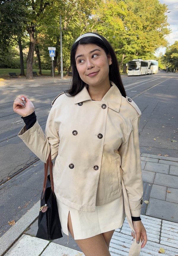 Une femme en trench beige se tient près d’une rue bordée d’arbres, avec des bus et des rails de tram derrière elle.