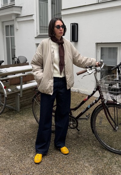 Persona con chaqueta beige, pantalones oscuros, zapatos amarillos y gafas de sol, de pie junto a una bicicleta con cesta delantera en un patio al aire libre.