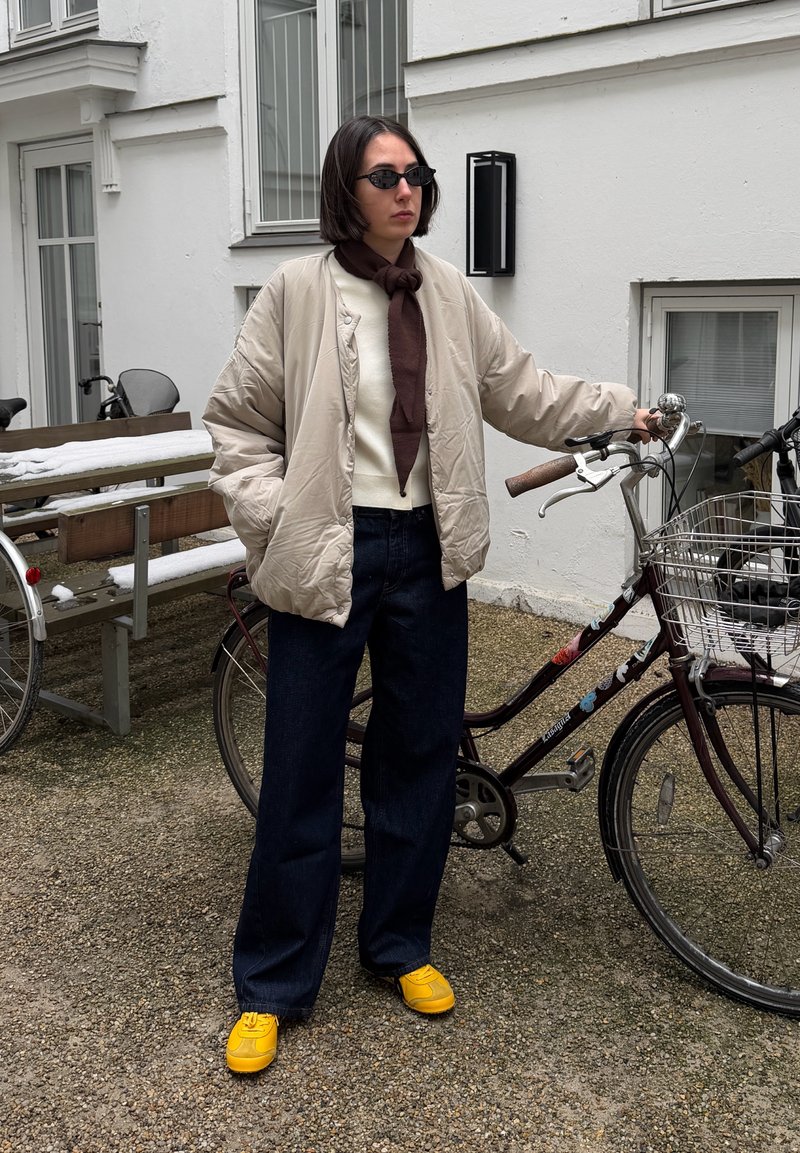 Personne portant une veste beige, un pantalon foncé, des chaussures jaunes et des lunettes de soleil, debout à côté d'un vélo avec un panier avant dans une cour extérieure.