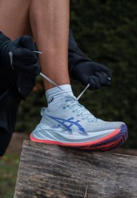 Világoskék futócipő lila akcentusokkal, texturált hálós felsőrész, fehér fűzők és piros talp. A fűzők éppen meg vannak kötve.