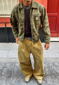 Olivengrønn kamuflasjedress med to frontlommmer, svart t-skjorte under, og løse beige bukser, kombinert med unike fottøy.