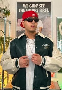 Grüne und cremefarbene Varsity-Jacke mit gerippten Bündchen, über einem weißen Hemd getragen. Rote Kappe und Sonnenbrille, übereinander getragene silberne Halsketten und Ringe an den Händen.