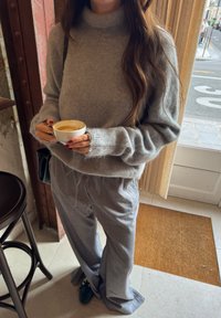 Grå, løstsiddende sweater med høj halsudskæring og blød tekstur, kombineret med lysegrå bukser. Holder en hvid kop med kaffe.