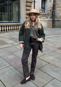 Grüne Feldjacke, braunes karierte Hemd, braune Hose, dunkle Slipper und ein beigefarbener breiter Hut, stehend auf steinernen Pflaster mit einem Gebäudehintergrund.