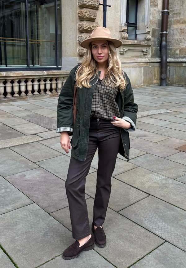 Veste de champ verte, chemise à carreaux marron, pantalons marron, mocassins noirs, et un chapeau à large bord beige, se tenant sur un pavé en pierre avec un bâtiment en arrière-plan.