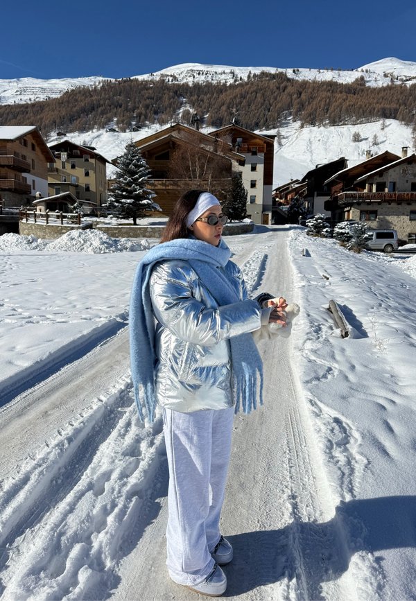 Doudoune argentée métallique, écharpe bleu clair, pantalon blanc et bottes grises. Paysage enneigé avec chalets en bois et montagnes en arrière-plan.