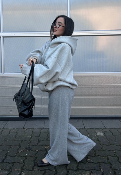 Mujer con sudadera gris claro de gran tamaño y pantalones de pierna ancha, sosteniendo un bolso negro, de pie sobre un suelo pavimentado frente a una puerta de garaje metálica.