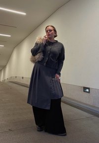 Grauer Wollcardigan mit Knöpfen, plissierter grauer Rock, schwarze weite Hose, flauschige beigefarbene Handtasche und silberne Ringe in einem modernen Flur.