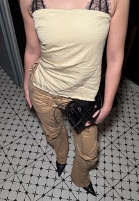 Personne portant un haut beige sans bretelles avec des lanières en dentelle noire, un pantalon marron froissé, des chaussures noires pointues, tenant un sac à main en cuir verni noir.