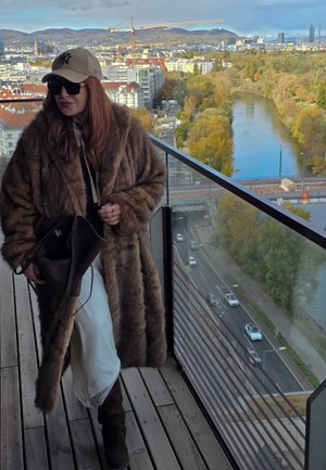Abrigo de piel sintética marrón, vestido blanco con encaje, sudadera negra, gafas de sol y gorra beige. Vista urbana panorámica con un río y árboles.