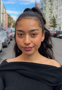 Czarna bluzka bez ramiączek z teksturowanej tkaniny. Długie, falowane czarne włosy. Naturalny makijaż z subtelnym kolorem ust. W tle widać samochody i budynki.